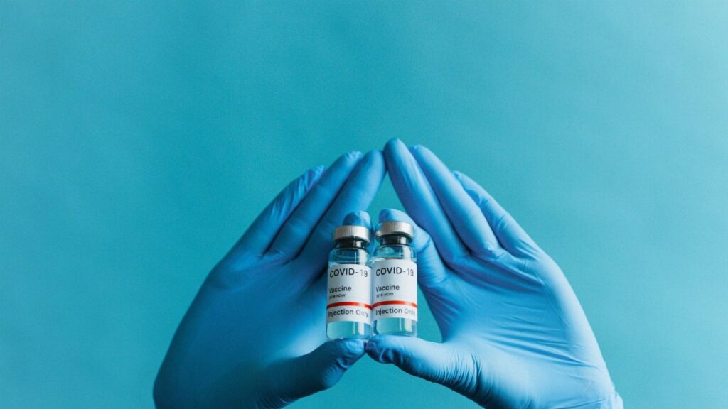 Blue gloved hands holding COVID-19 vaccine vials on a blue background.