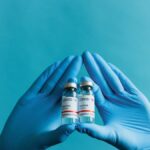 Blue gloved hands holding COVID-19 vaccine vials on a blue background.