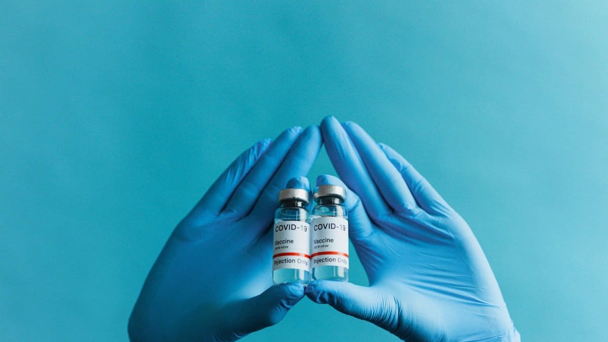 Blue gloved hands holding COVID-19 vaccine vials on a blue background.