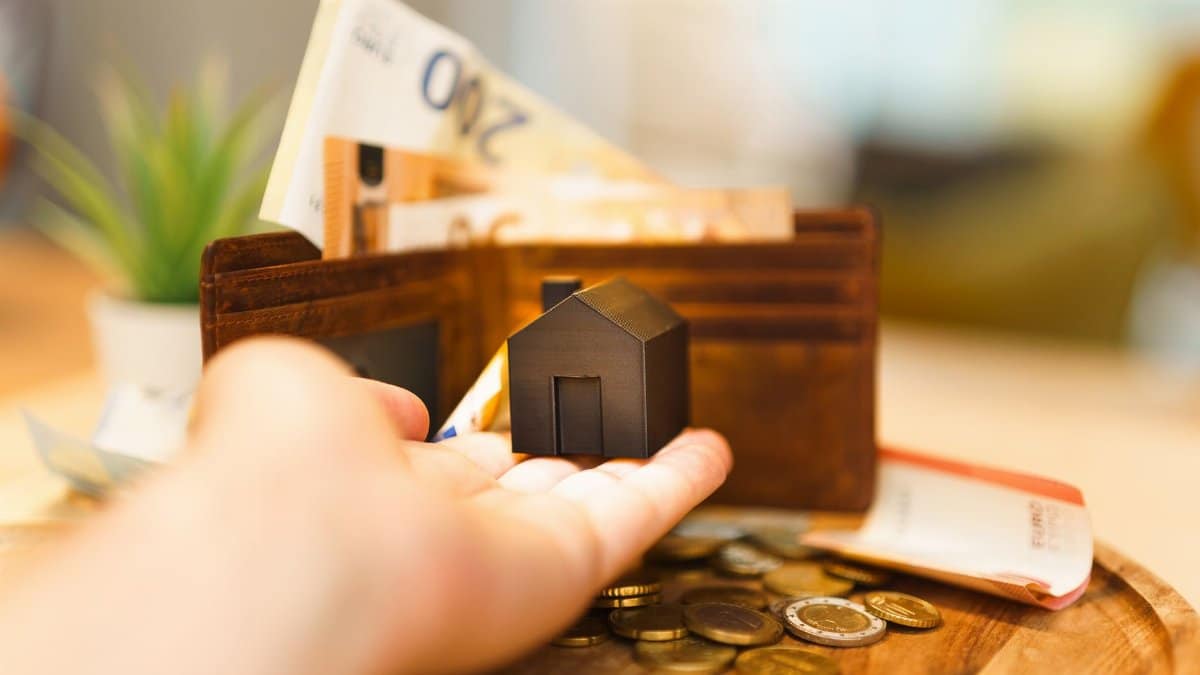 A hand holding a small house model with euro notes and coins nearby, illustrating real estate investment and finance.