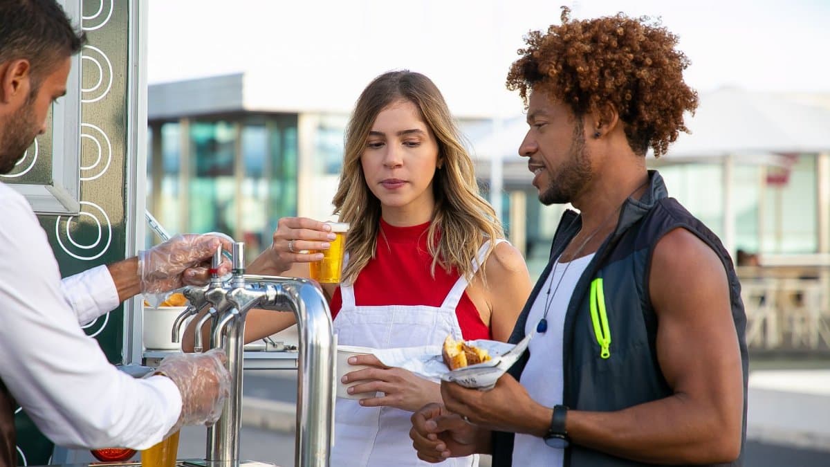 Salesman in gloves pouring alcohol drink from beer tap to multiethnic couple on street cafe