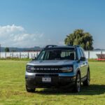 A silver Ford Bronco park on a grassy field under a blue sky, showcasing a rural landscape.
