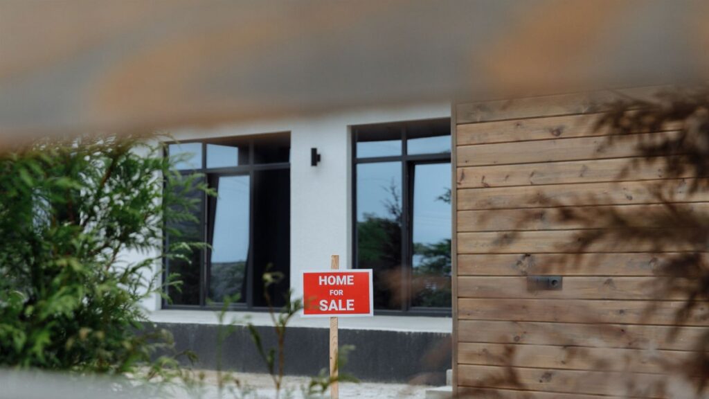 A contemporary home with a 'For Sale' sign in the front yard, framed by greenery.