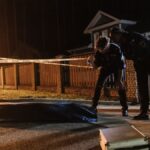 Police officers investigate a crime scene at night, examining evidence near a residential area.