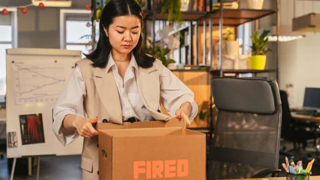 An Asian woman packs a box labeled 'FIRED', symbolizing job loss in an office setting.