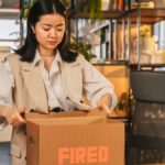 An Asian woman packs a box labeled 'FIRED', symbolizing job loss in an office setting.