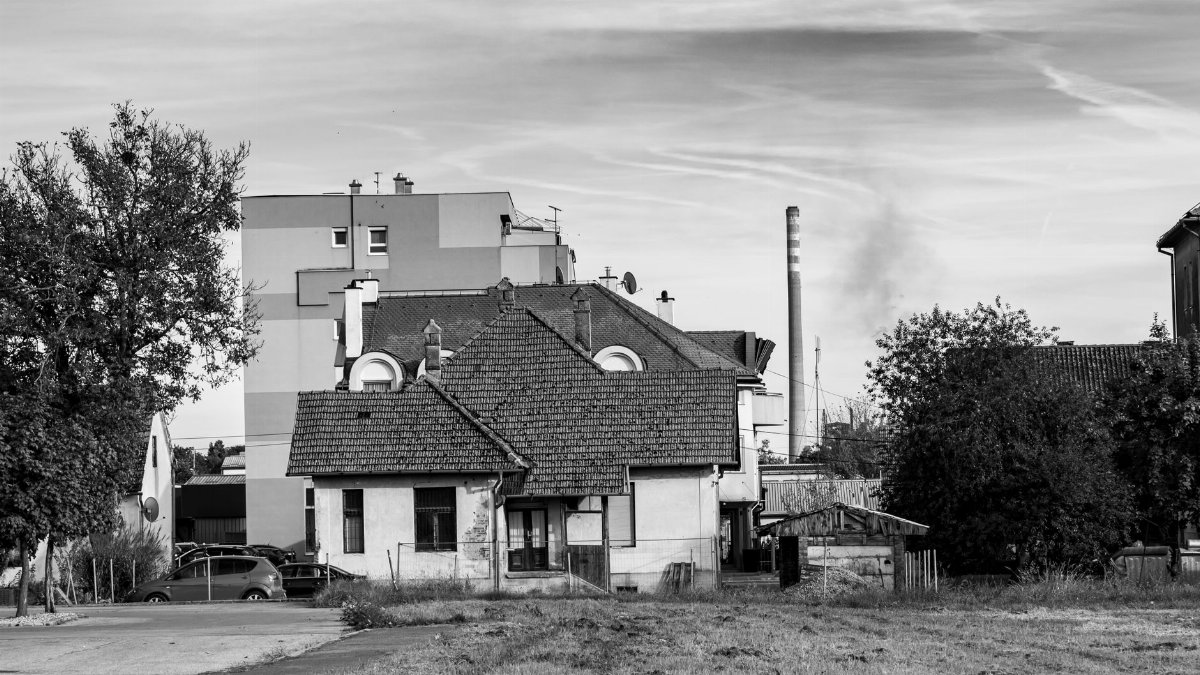Black and white photo of a rural house with industrial structures in the background, Kutina, Croatia.