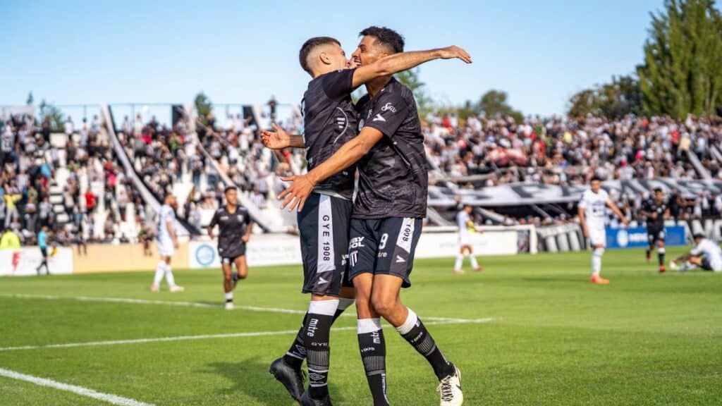 Two soccer players in black jerseys celebrate a goal on a sunny day at a packed stadium.