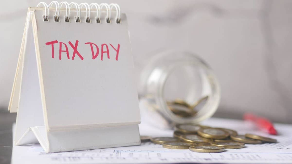A close-up of a table calendar marked 'Tax Day' with a jar of coins and notes, symbolizing financial planning.