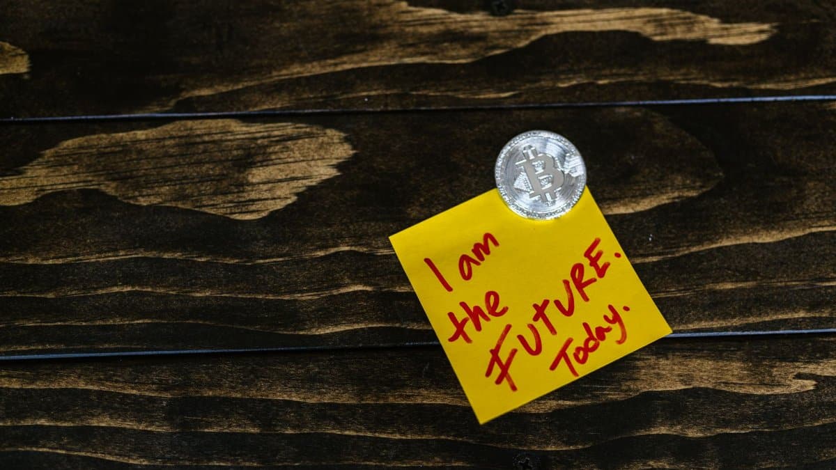 Bitcoin coin pinned on a wooden table with a yellow sticky note that reads 'I am the FUTURE. Today.'