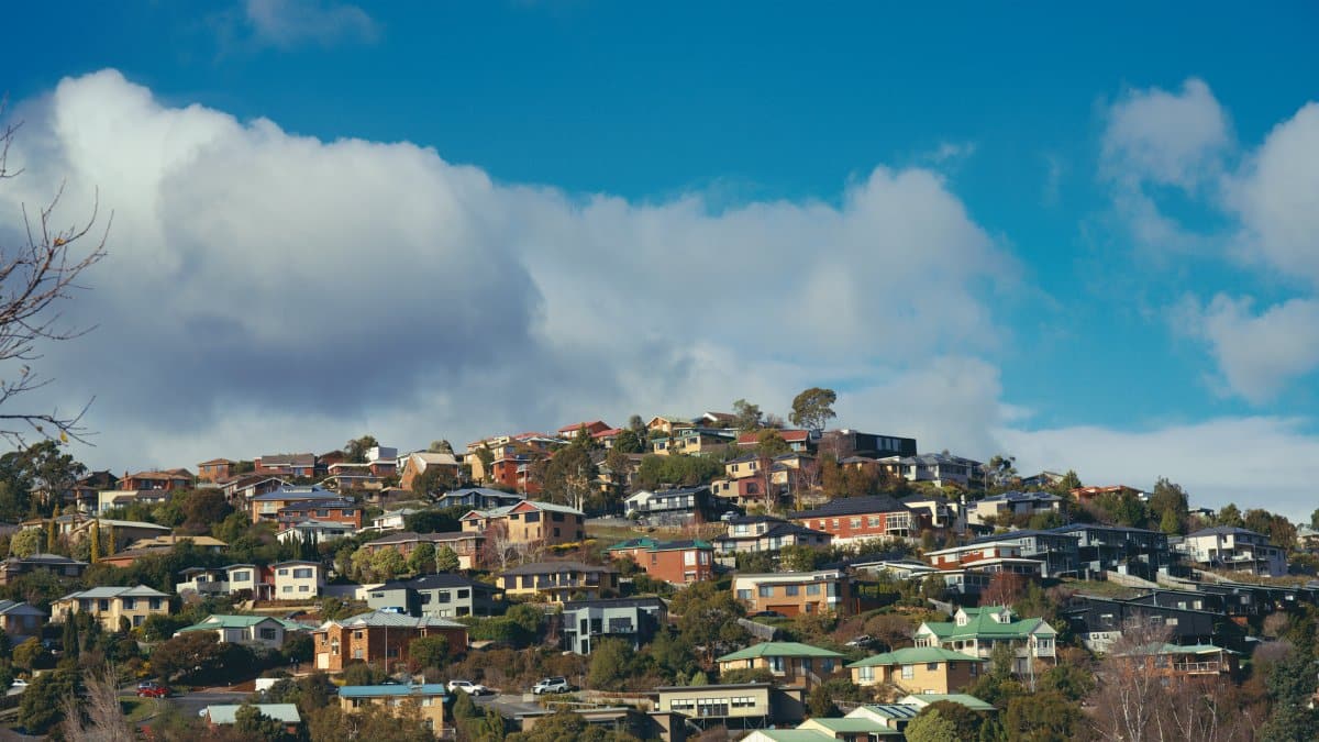 Hillside view of residential houses beneath a clear, bright sky with fluffy clouds.