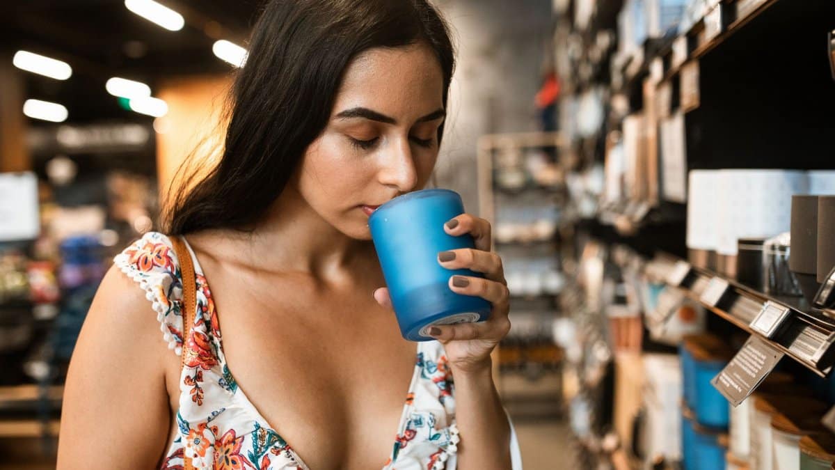 A woman in a floral dress smells a blue scented candle in a store aisle, savoring its fragrance.