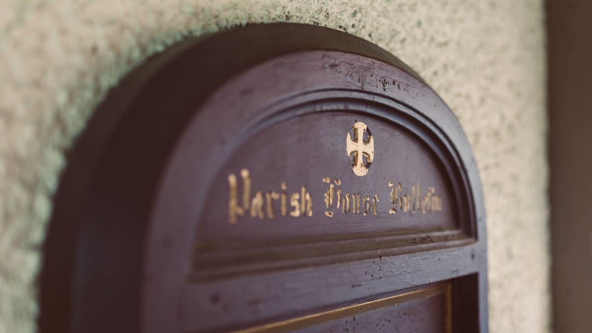 Close-up of a wooden Parish Notice Bulletin board with a cross emblem, in a church's entryway.