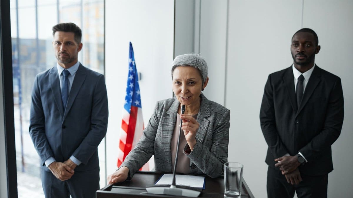 A female politician delivers a speech with bodyguards and an American flag in the background.