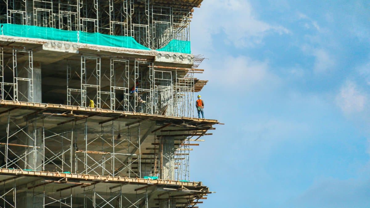 A construction worker stands on a scaffolding at a high-rise building under a blue sky.