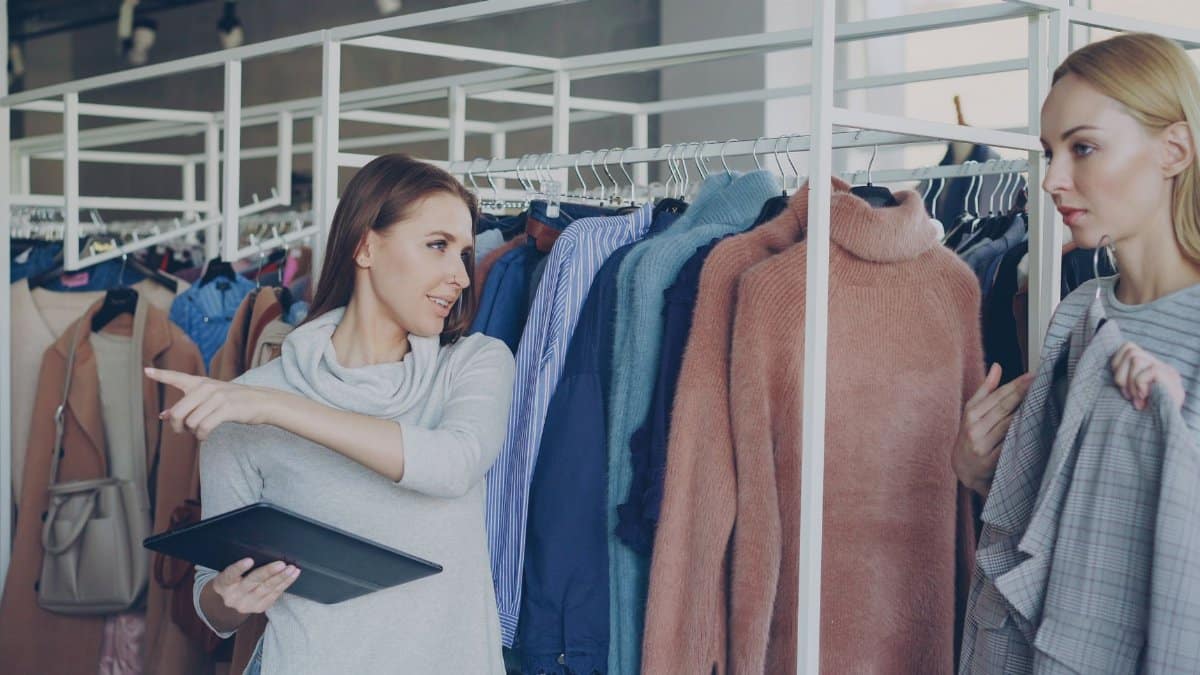 Two women shopping for clothes, one holding a tablet, discussing options in a store.