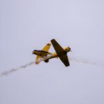 A vintage yellow aircraft soaring through the sky leaving a trail of smoke during a daytime air show.