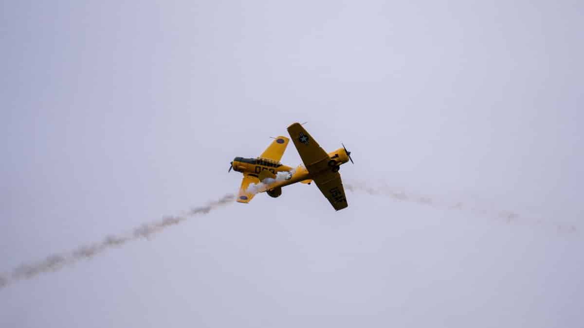 A vintage yellow aircraft soaring through the sky leaving a trail of smoke during a daytime air show.