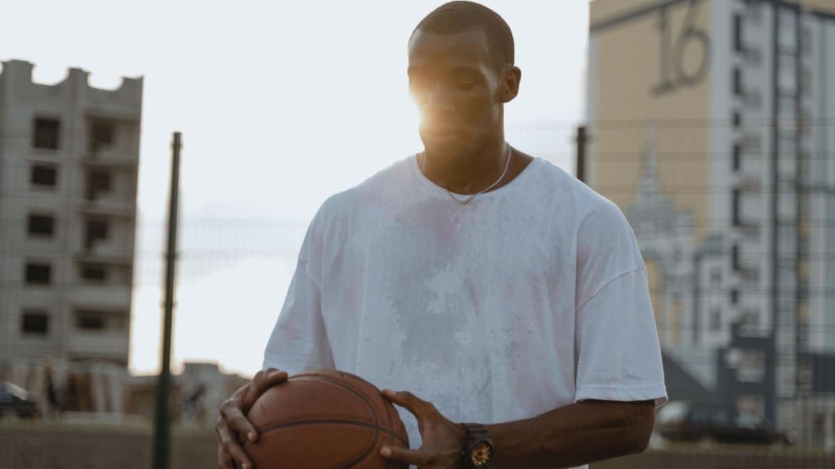 A man holding a basketball outdoors with the sun setting behind, creating a dramatic urban scene.