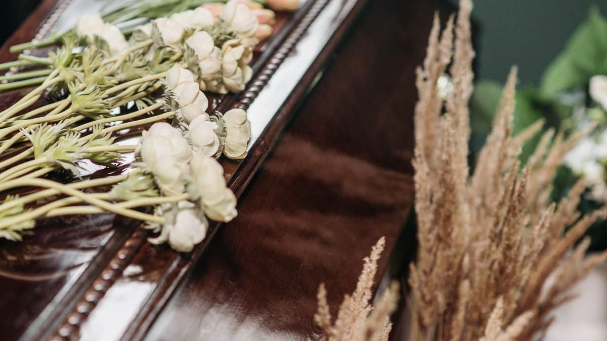 A solemn funeral arrangement featuring a wooden casket adorned with white ranunculus and dried grasses.