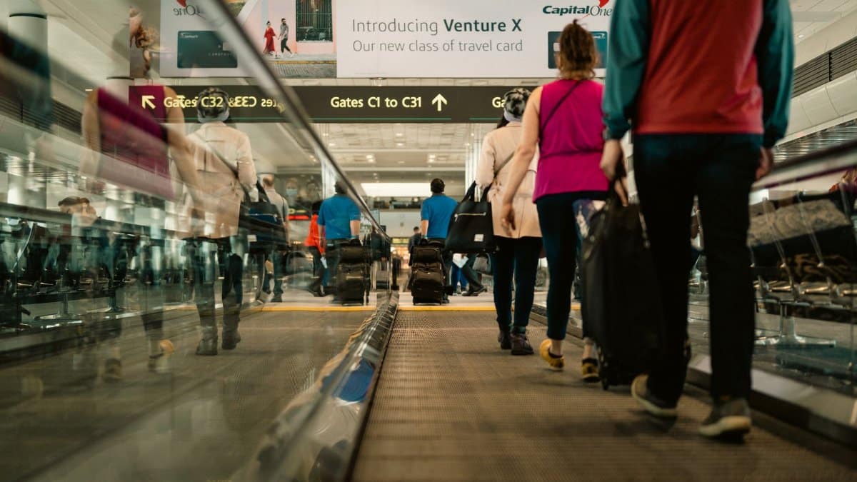 Passengers walking on a moving walkway in a busy airport terminal with signs for gates.