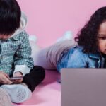 Little multiethnic kids in casual outfit pastime on floor and using computer and smartphone on pink background