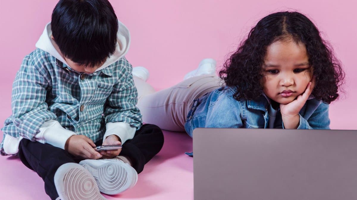 Little multiethnic kids in casual outfit pastime on floor and using computer and smartphone on pink background