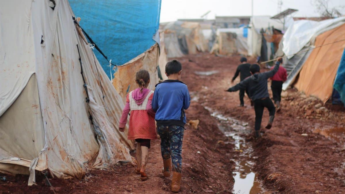Back view group of kids strolling on dirty ground with water between worn out tents in poor district of small settlement