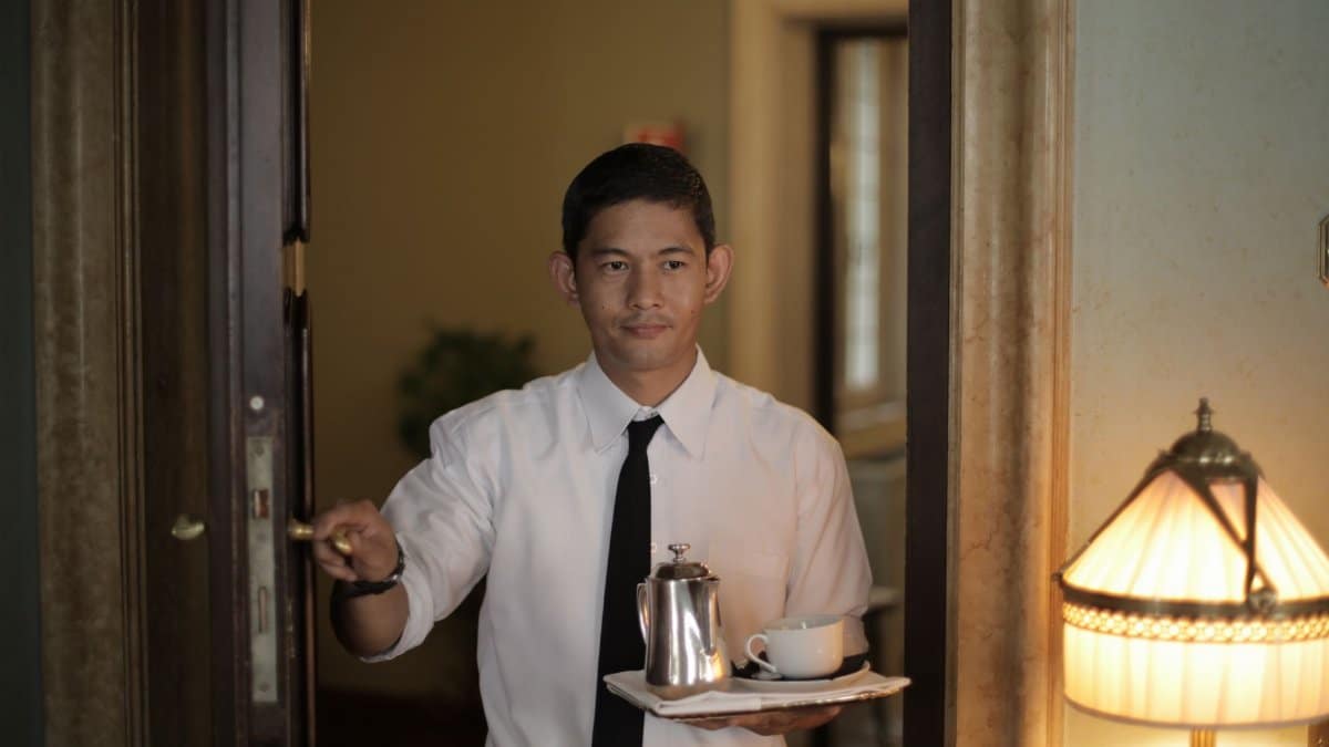 Positive ethnic male employee carrying tray with coffee while serving guest in cozy hotel room
