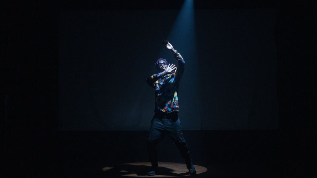 Dynamic image of a stylish performer dancing under a spotlight on stage, showcasing fashion and movement.