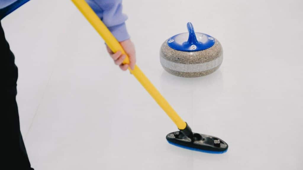Crop anonymous sportsman with special broom sweeping ice sheet in front of sliding curling granite stone while playing sportive game