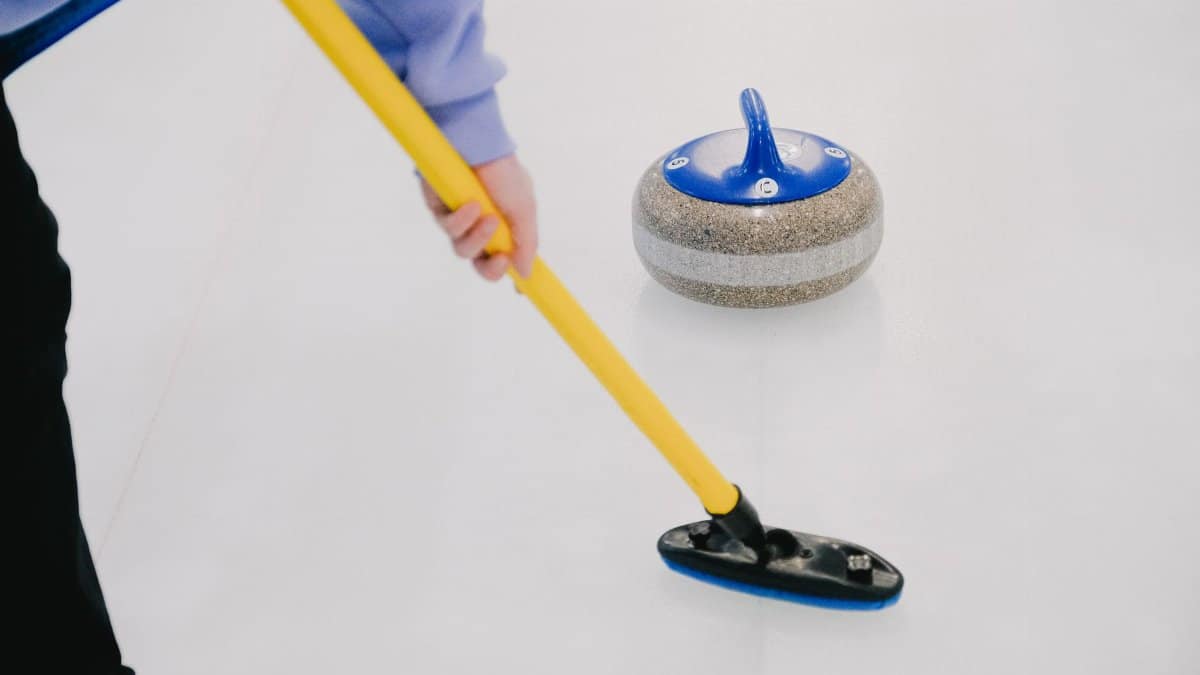 Crop anonymous sportsman with special broom sweeping ice sheet in front of sliding curling granite stone while playing sportive game
