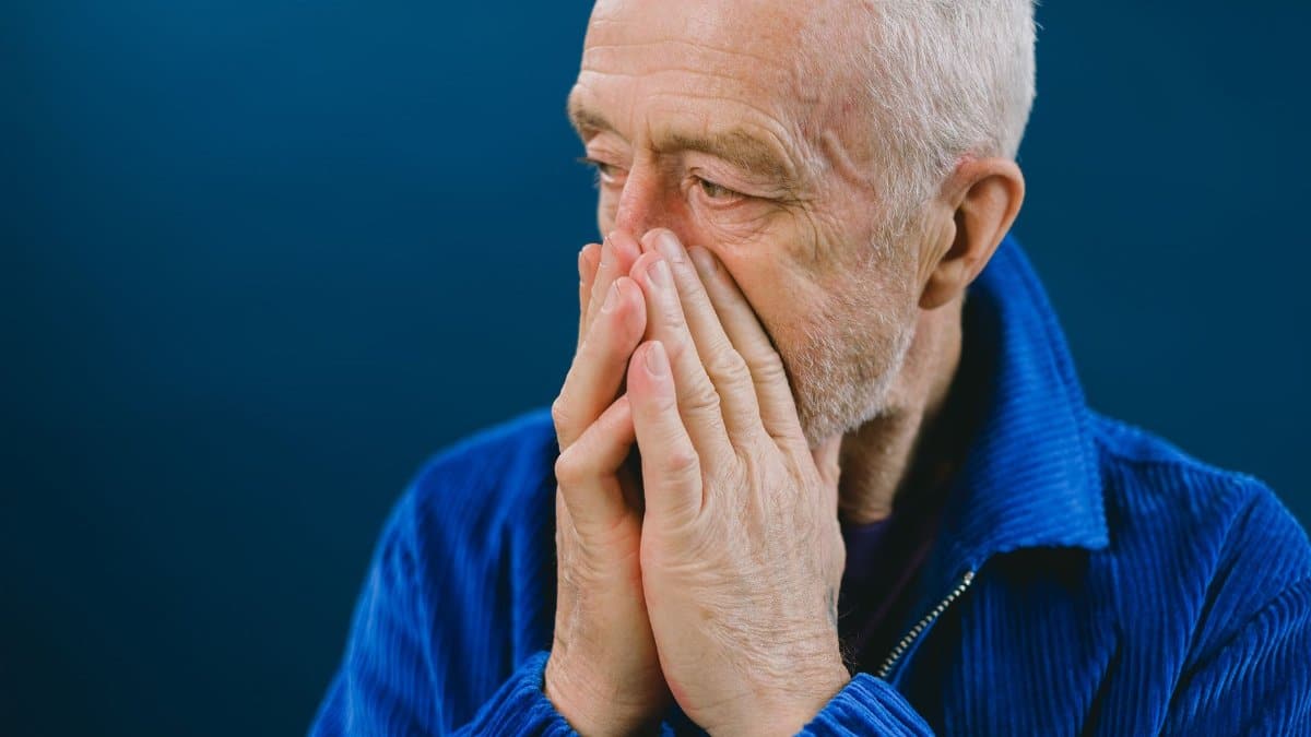 Serious anxious bearded senior gray haired male in blue jacket covering mouth with hands while looking away against blue background