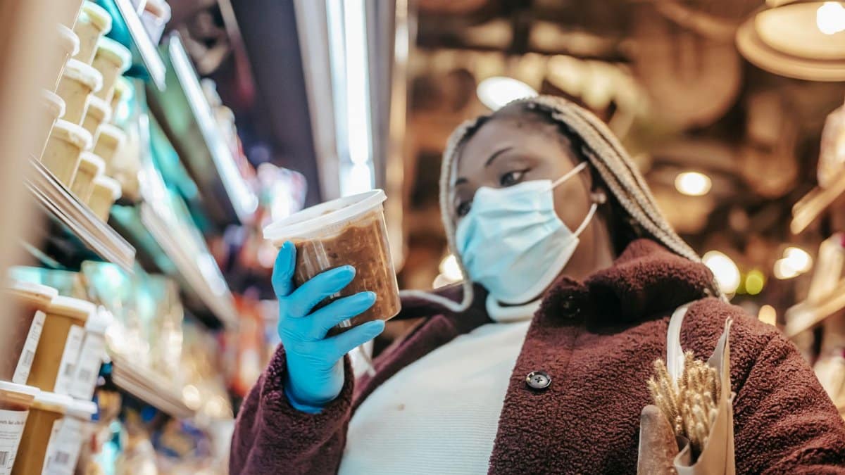 Low angle of African American female in mask and latex gloves considering food while shopping in supermarket during pandemic of coronavirus