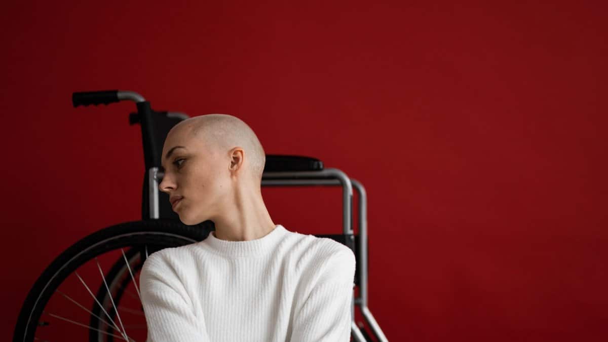 Thoughtful young bald female with disability in warm sweater leaning on wheelchair and looking away against red background