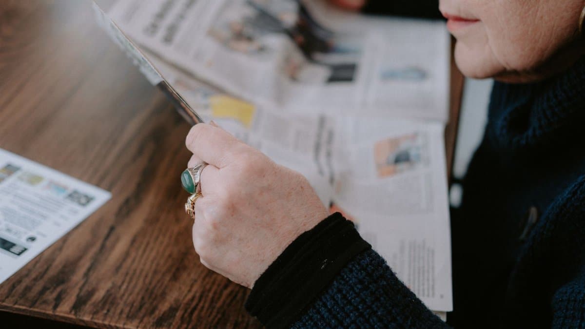 A woman reads a newspaper at a wooden table, focusing on leisure and information.