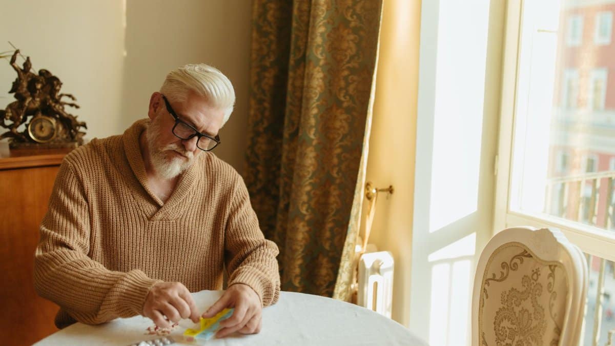 Senior man with gray hair organizing pills at a table by a window, emphasizing daily health routine.