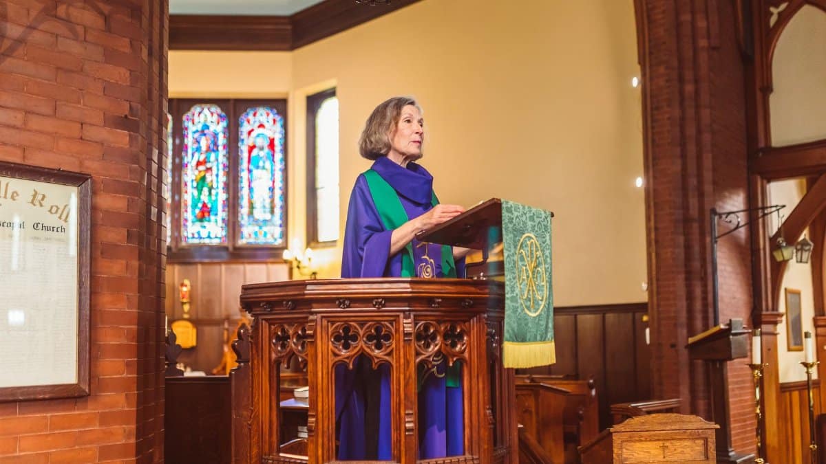 Elderly woman in cassock delivering sermon from ornate podium inside a historic church.