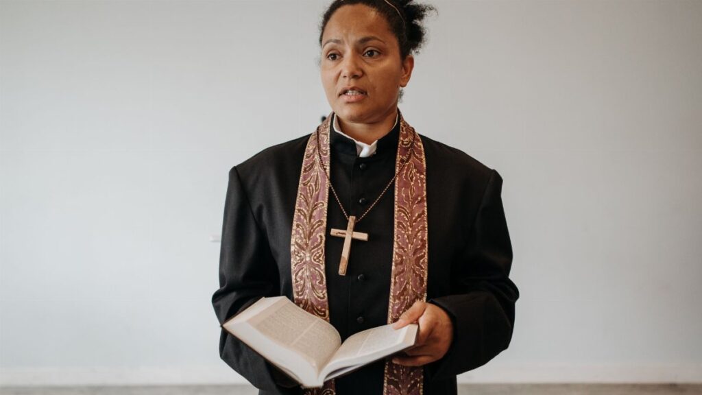A female pastor in clerical attire stands with an open Bible in a church setting.