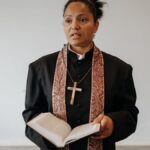 A female pastor in clerical attire stands with an open Bible in a church setting.