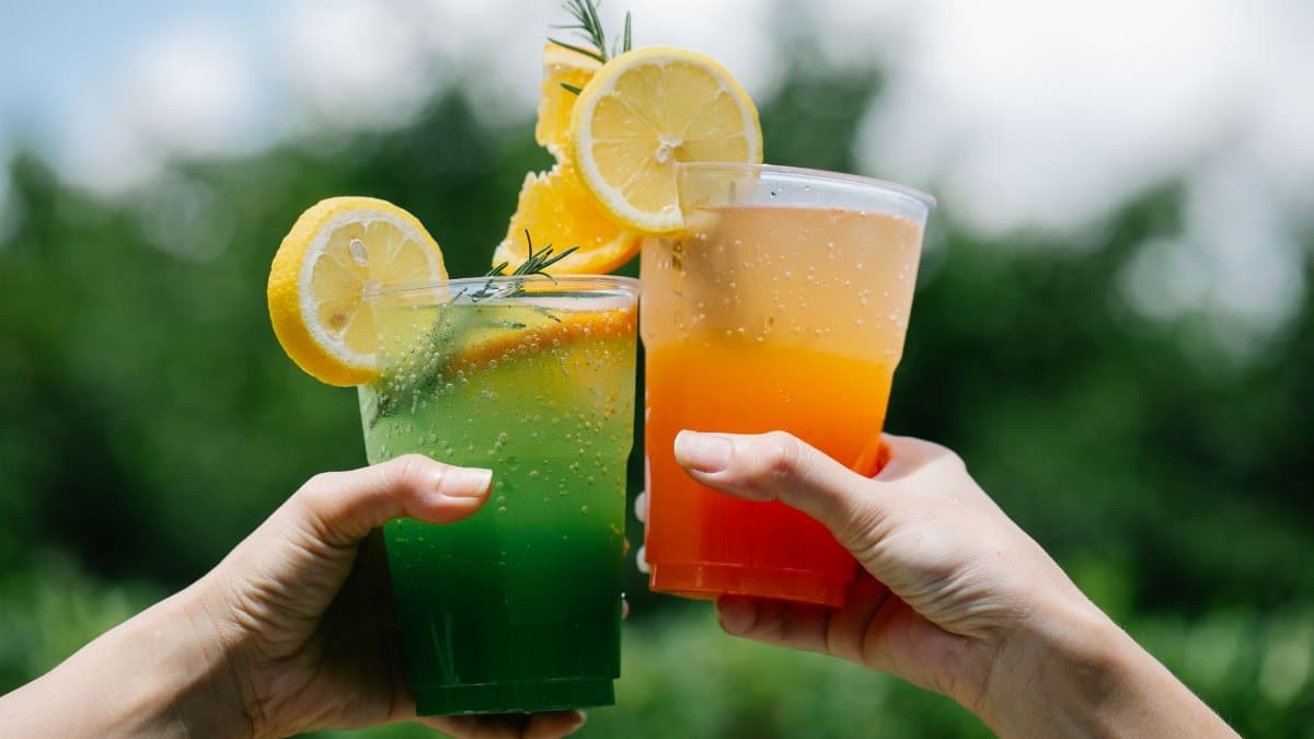 Two hands holding colorful citrus lemonades in a vibrant summer outdoor setting.