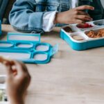 Crop faceless classmates sitting at table with lunch boxes with various food and eating during break