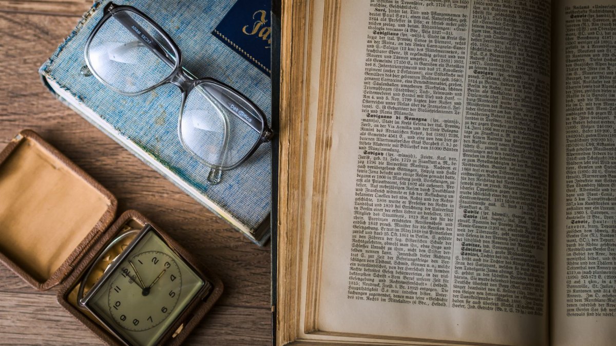 A vintage setting with antique books, watch, and reading glasses on a wooden table.