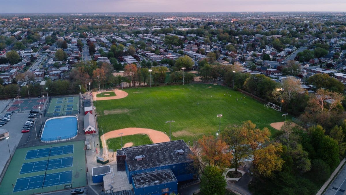 Drone shot of a vibrant sports complex and surrounding neighborhood in Philadelphia during sunset, showcasing a baseball field, tennis courts, and pool.