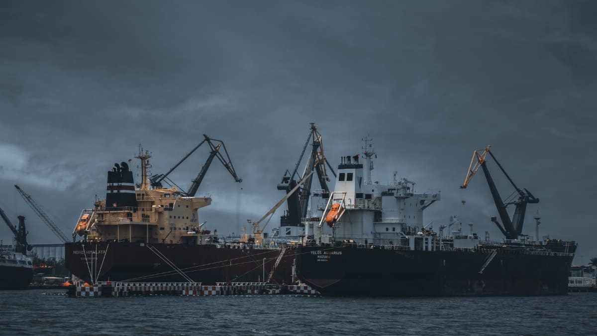 Cargo ships at a bustling port under cloudy skies, cranes ready for loading.