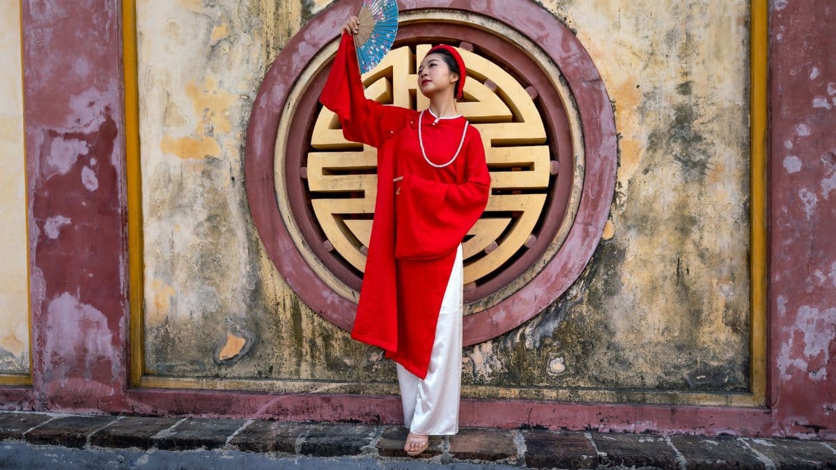 Asian woman wearing traditional clothing and holding a fan, posing against a decorative background.