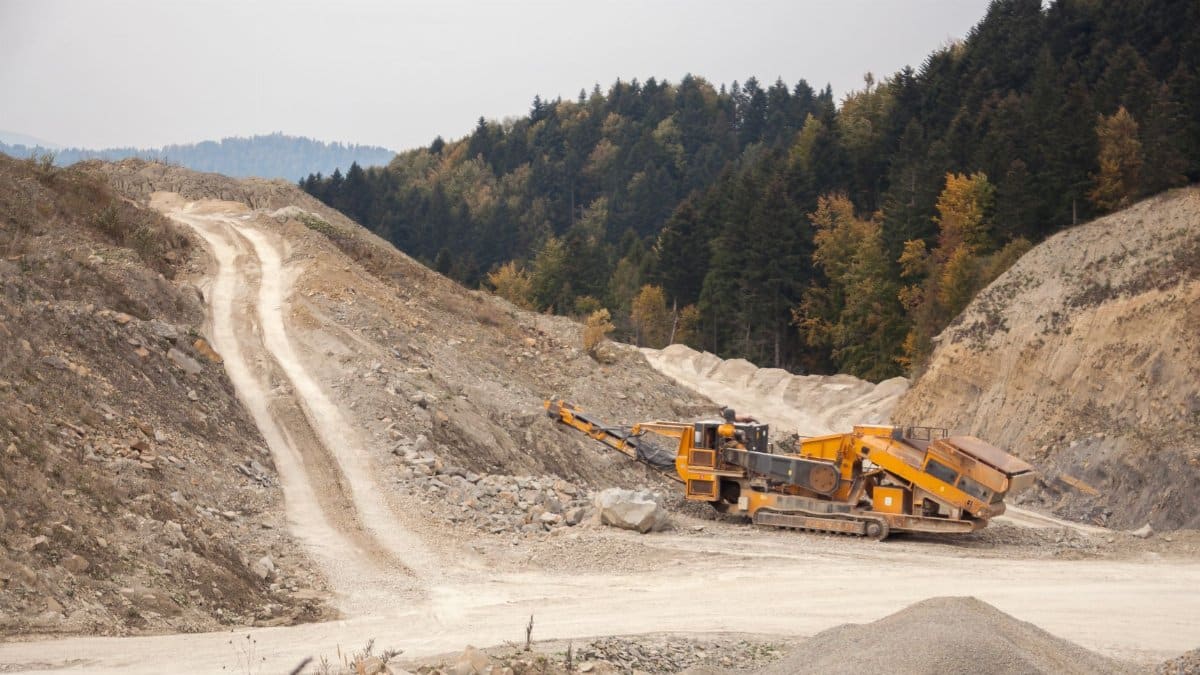 Industrial crusher machine reducing rocks near big pile of soil with tracks in nature near woods with lush green trees