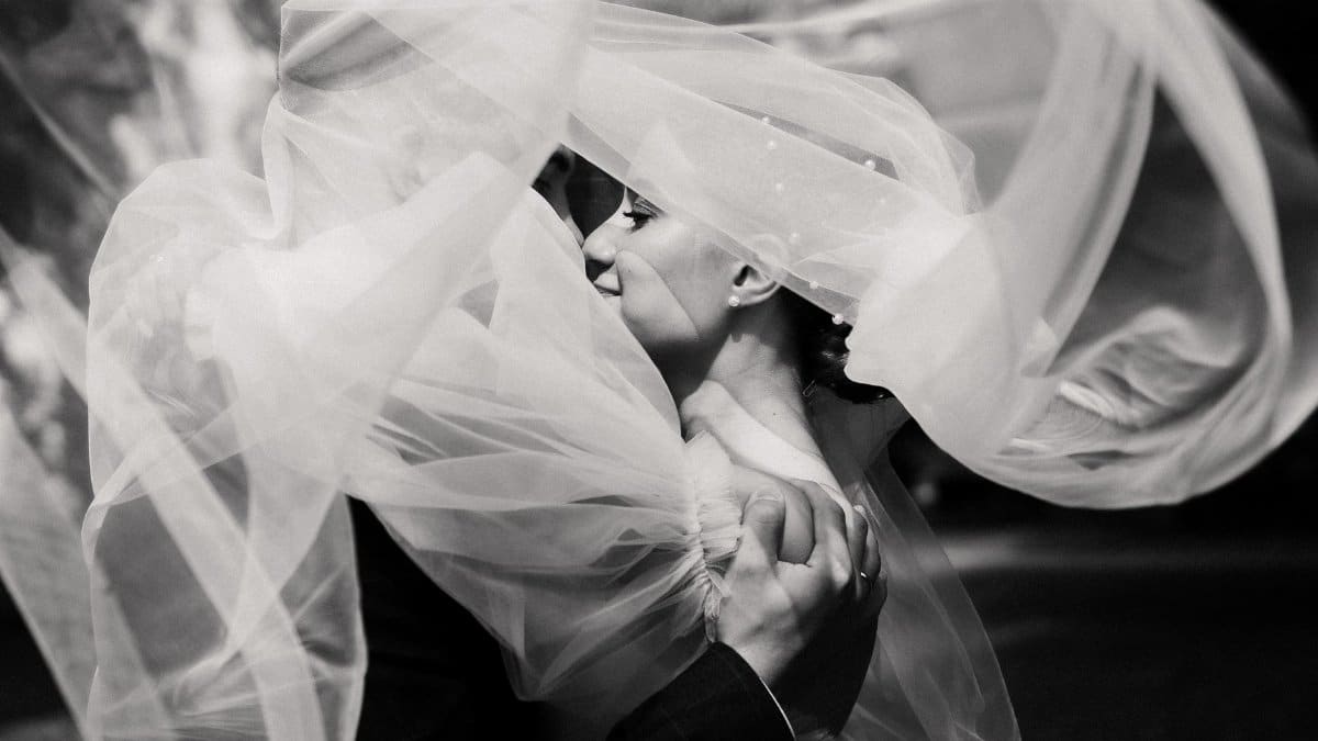 Bride and groom share a tender kiss beneath a flowing veil in elegant grayscale.