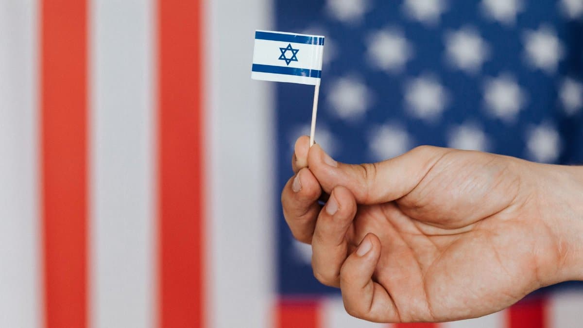 Closeup of crop unrecognizable person holding small flag of Israel before huge flag of United States of America on background