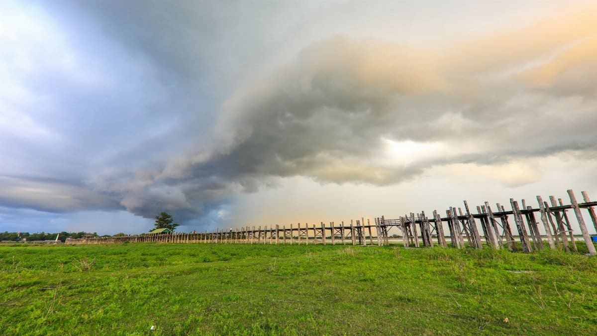 A dramatic overcast sky over U Bein Bridge in Amarapura, showcasing vibrant green fields.
